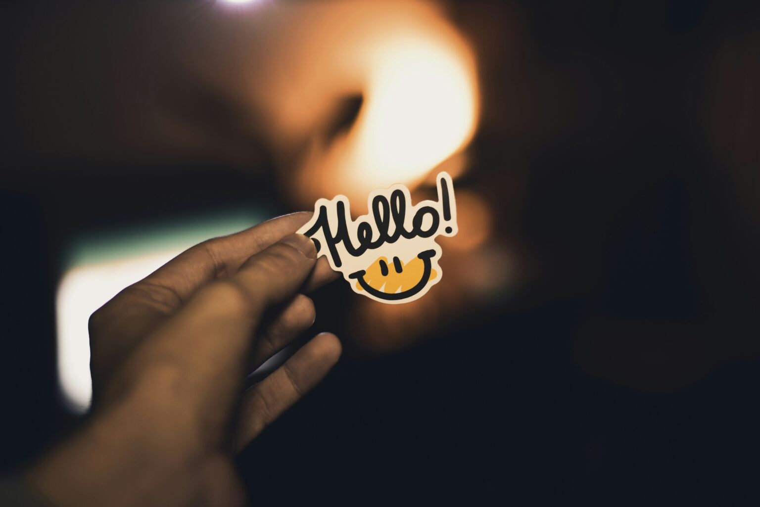 A hand holding up a smiling 'Hello!' sticker against a warm, glowing background, representing a website that welcomes visitors in